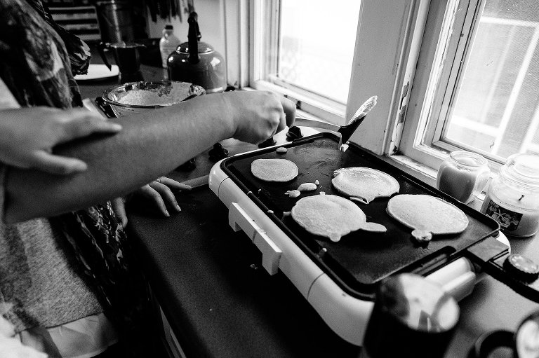 Black and white. Daughter holds onto mom's arm as she flips pancakes. 