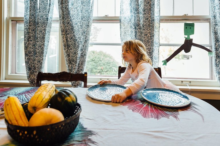 Girl sets the breakfast table. A construction paper "frankenstein" decoration is in the window behind her.