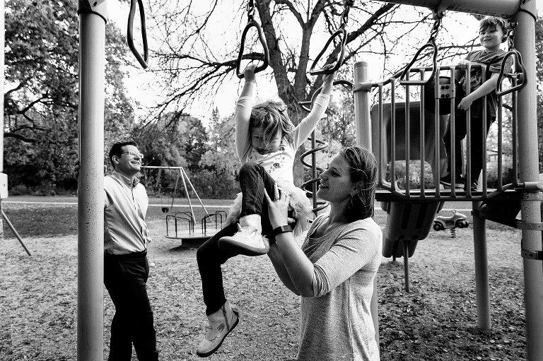 Mom helps girl across monkey bars at park while big brother smiles at them. Dad smiles at the young boy. Black and white. 