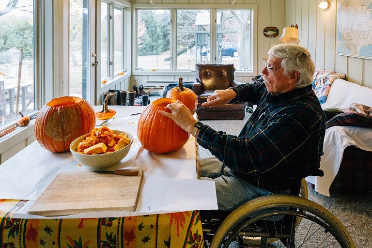 Grandpa carves pumpkin at a table. 3 pumpkins on the table. 