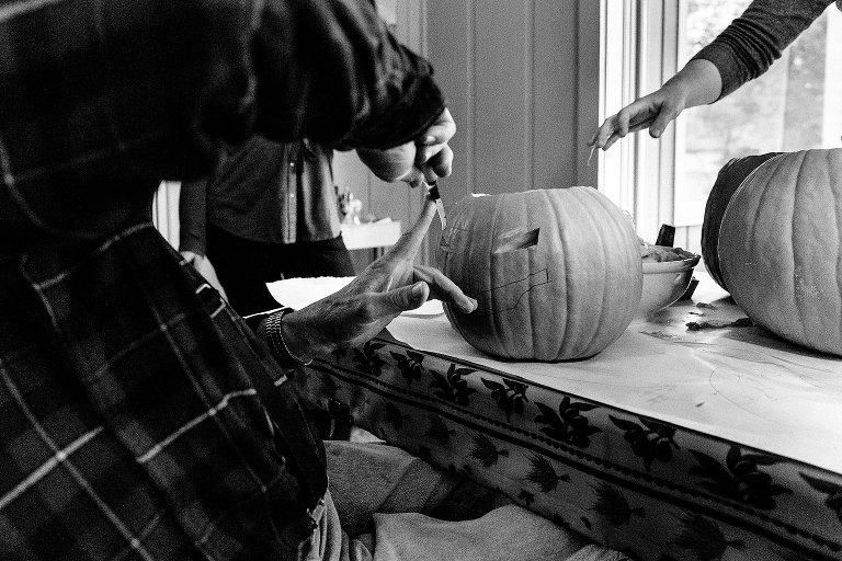 Black and white. Grandpa carefully places his finger against the blade of a knife while carving a pumpkin. A hand enters the frame at the top to dump more pumpkin guts. 