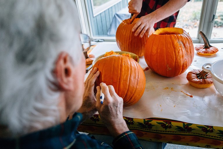 Grandpa draws on pumpkin while another person works on carving out the insides of a different pumpkin across the table. 