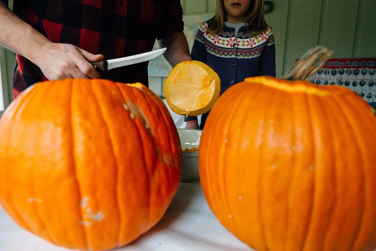 Man cuts off top of pumpkin with knife. Framed by two large pumpkins. 