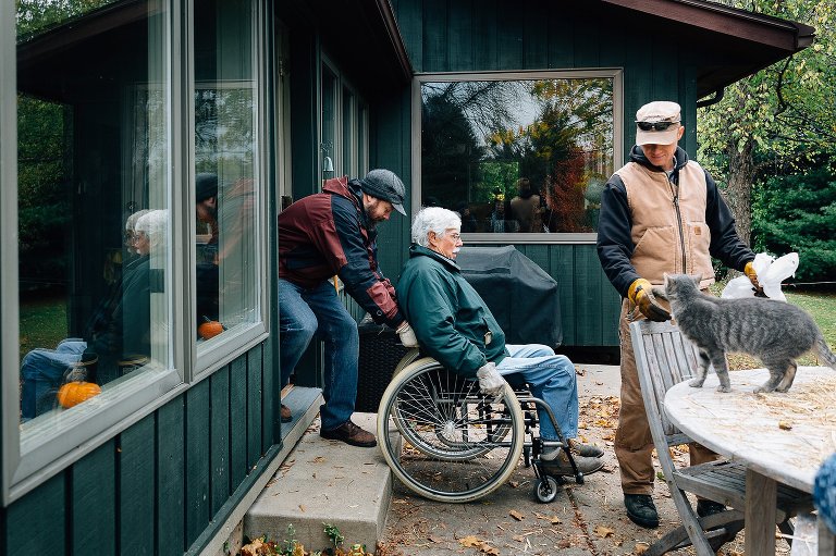 Man helps grandfather in wheelchair into the house. Man stands outside and pets farm cat. 