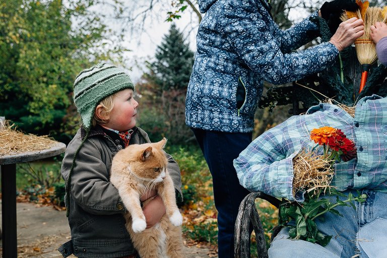 Preschool boy carries a farm cat and watches grandma and sister put finishing touches on scarecrow. 