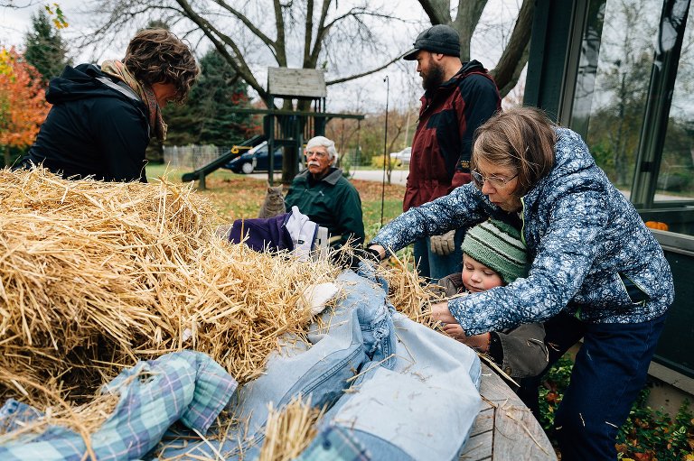 Family works together to make a scarecrow outside. There is a straw all over the table while grandma, mom, and grandson work on it. 