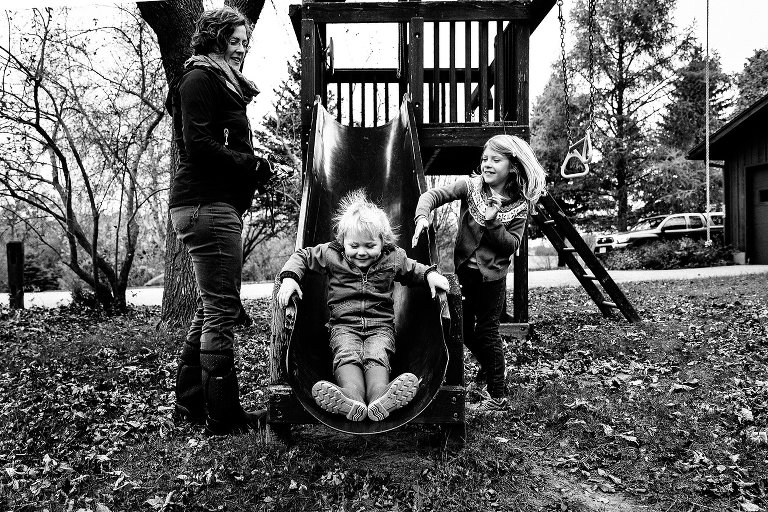Black and white. Boy comes down slide with static in hair. A girl helps push him to go faster as his mother watches. 