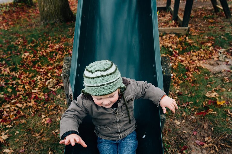 Toddler boy comes down a green slide in the fall. 