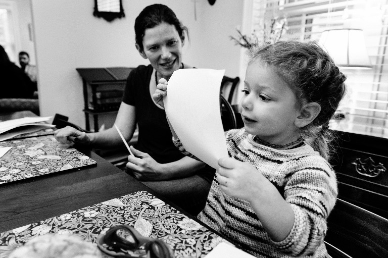 Black and white. Mother sits at dining room table while preschool child shares a story she wrote.