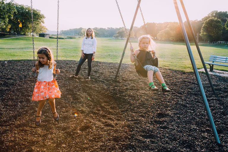 Two girls swing at the park. Mother pushes the youngest girl. Sun flare enters the frame. 