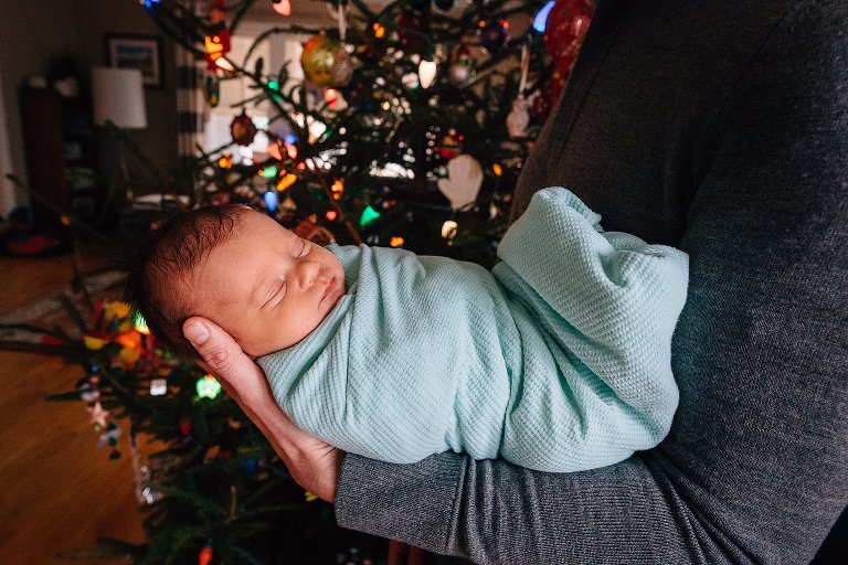 Dad holds sleeping newborn in arms. Christmas tree in background. 