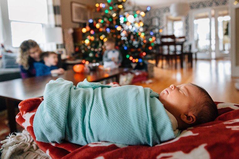 Sleeping newborn lies in foreground in focus while mother and two older boys play together in background near Christmas tree. 
