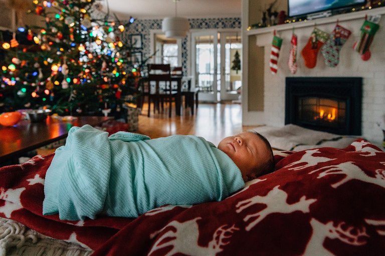 Newborn lies on reindeer blanket with Christmas tree and fire with stocking in the background. 