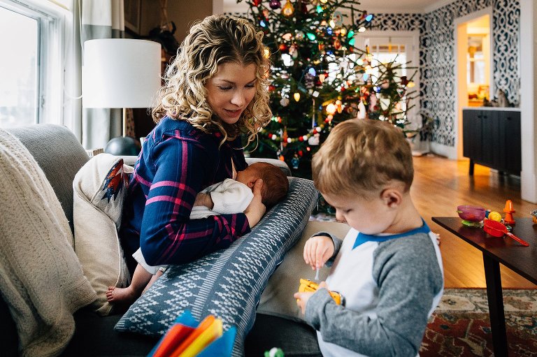 Mother nurses newborn while engaged iwht preschool son. Christmas tree in background. 