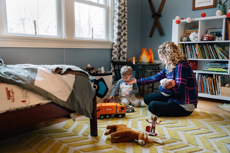 Mother reaches to gently touch preschooler's chin while holding newborn baby in one arm. they are sitting on bedroom floor. 