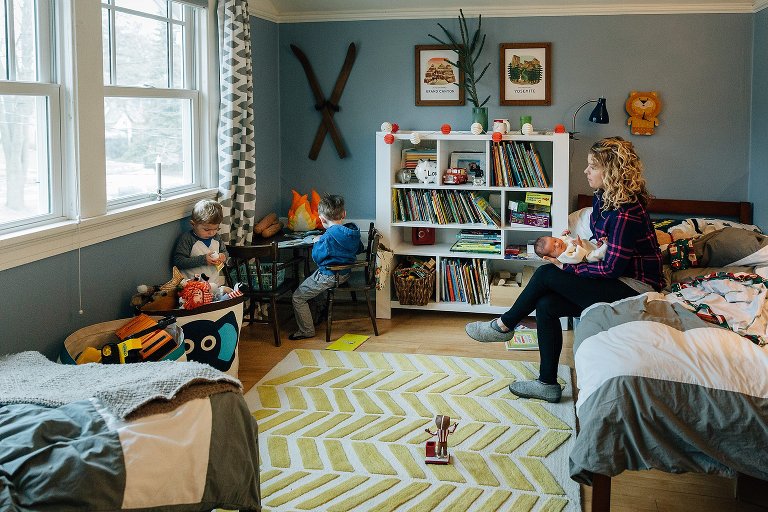 Mother sits in her children's room while they play. She holds newborn child on hte bed. 