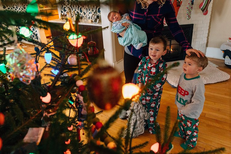 Christmas tree in foreground, two small boys wearing Christmas pajamas looking at tree in wonder while mother holds newborn in her right hand and touches the head of one boy with another. 