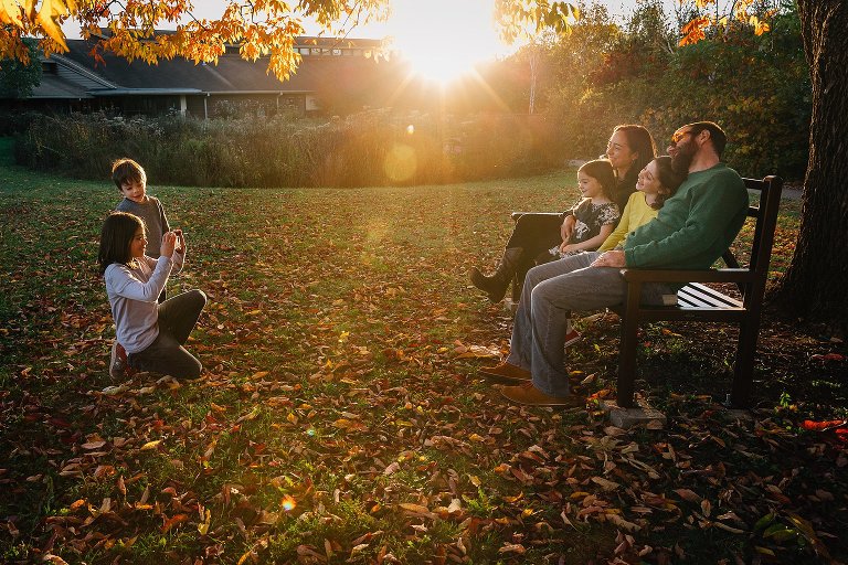 Two kids take pictures of family sitting on park bench with colorful fall leaves all around them.