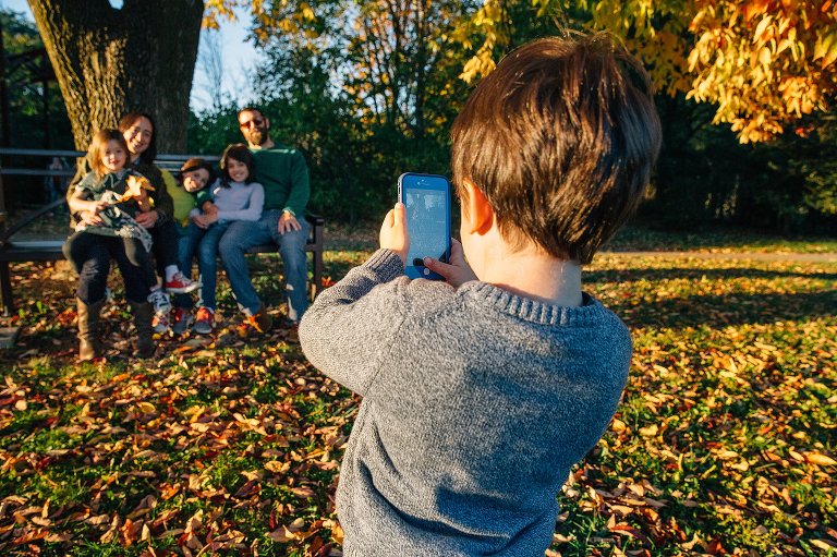 Little boy holds up smartphone to take a picture of family sitting on park bench with colorful fall leaves all around them.