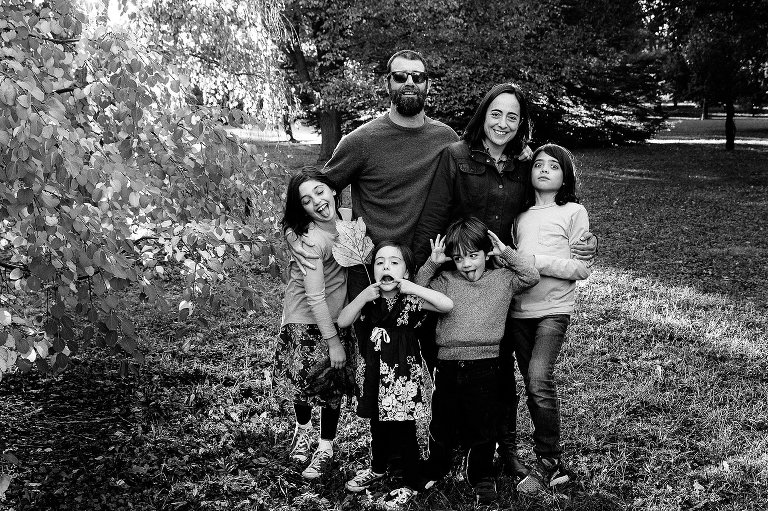 Black and white. Mom, Dad, and four kids stand for goofy family photo outside with trees behind them.