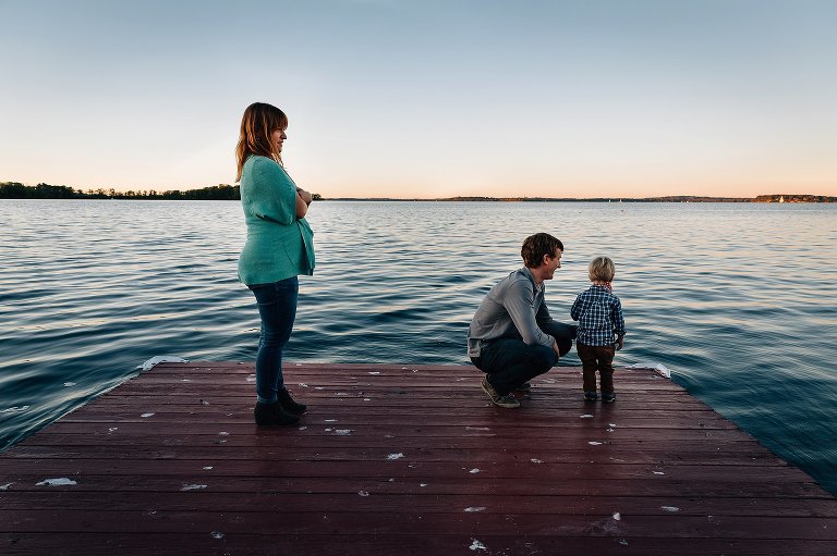 Pregnant mother standing on dock over a lake, father kneeling next to son.