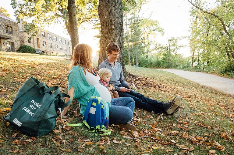 Pregnant mother, father, and son sitting on a grassy hill eating snacks.