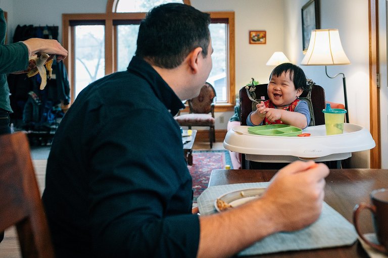Father eating breakfast at kitchen table watches little boy laugh and eat in highchair.