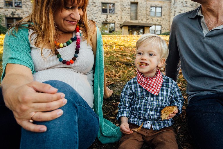 Pregnant mother, father, and son sitting on grass and leaves eating snacks.