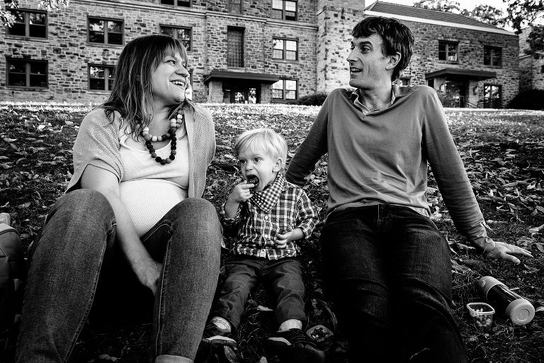 Black and white. Pregnant mother, father, and son sitting outside on the grass eating snacks.