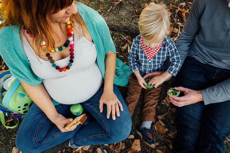 Pregnant mother, father, and son sitting on grass and leaves eating snacks.