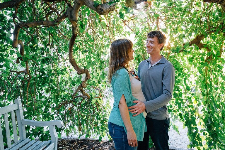 Pregnant mother and father stand under tree next to bench.