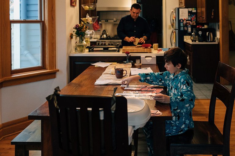 Young boy sits at dinning table playing cards while father cooks in the kitchen.