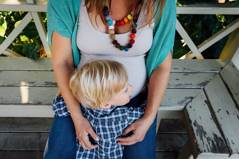 Pregnant mother sits outside on wood bench with little boy hugging her belly.