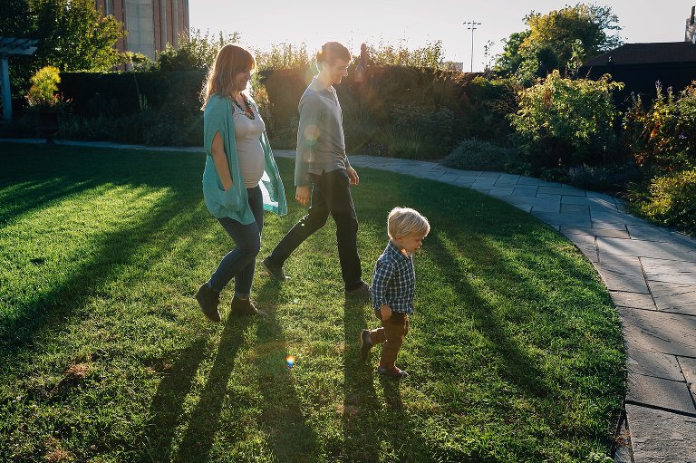 Pregnant mother, father, and son walk through the grass next to path and garden. Sun setting behind them.