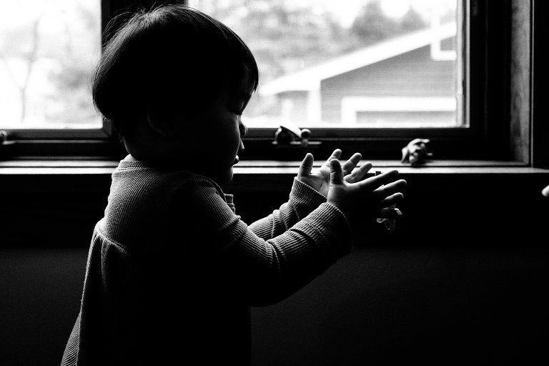 Black and white. Little boy looking at hands while standing next to window.