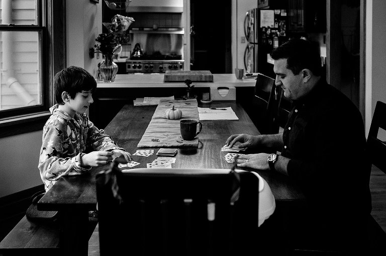 Black and white. Father and son sit at dinning table playing a game of cards.