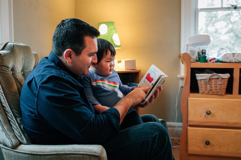 Father sitting in rocking chair reads book to young son sitting on his lap.