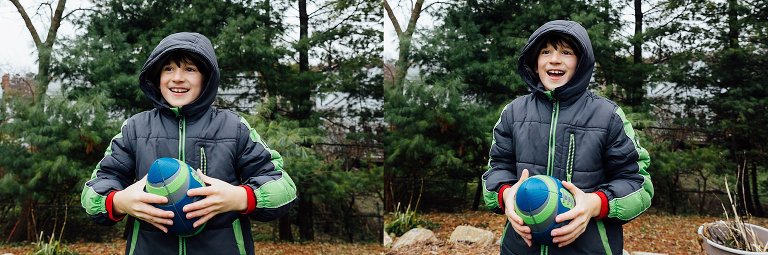 Young boy wearing coat and hood playing football in backyard.