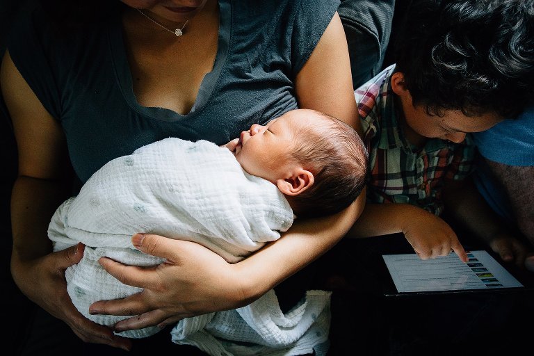 Mother sits on couch holding newborn baby next to son reading picture book.