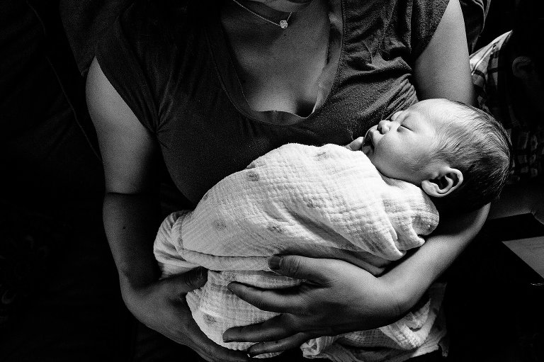 Black and white. Mother sits on couch holding sleeping newborn baby.