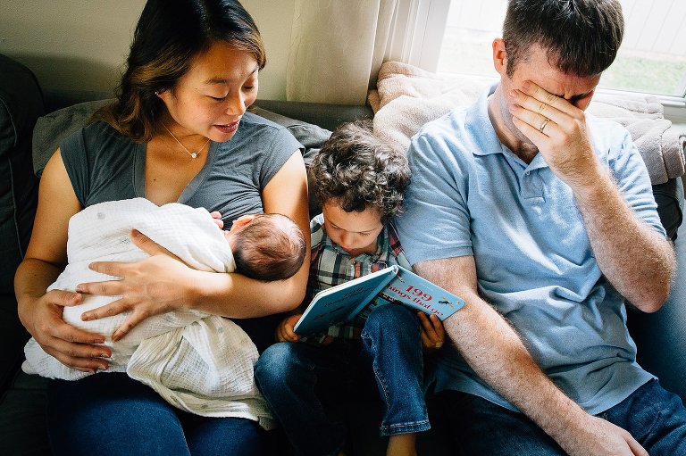 Mother holding newborn baby, father, and son sit on the couch reading a book.