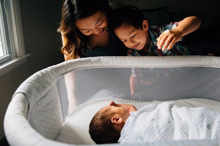 Mother and son look at newborn baby sleeping in bassinet.