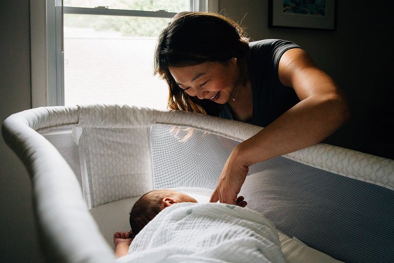 Mother smiles and touches newborn baby's face as he sleeps in baby bassinet.