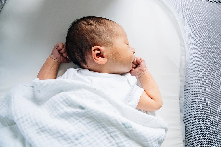 Newborn baby wearing white onesie with a white blanket sleeps in white baby bassinet.