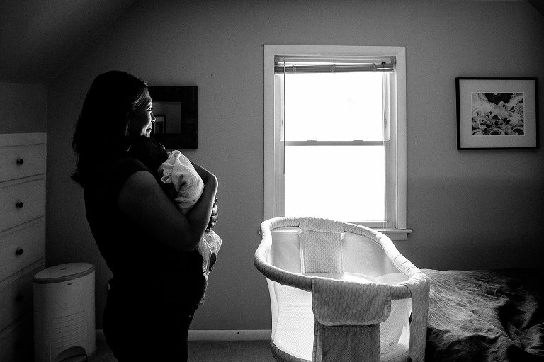 Black and white. Mother holds sleeping newborn baby as she stands next to baby crib.