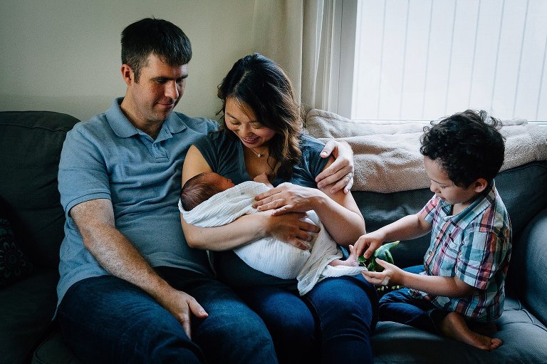 Mother holds swaddled newborn baby boy, Father sits next to them, little brother looks at baby's feet.