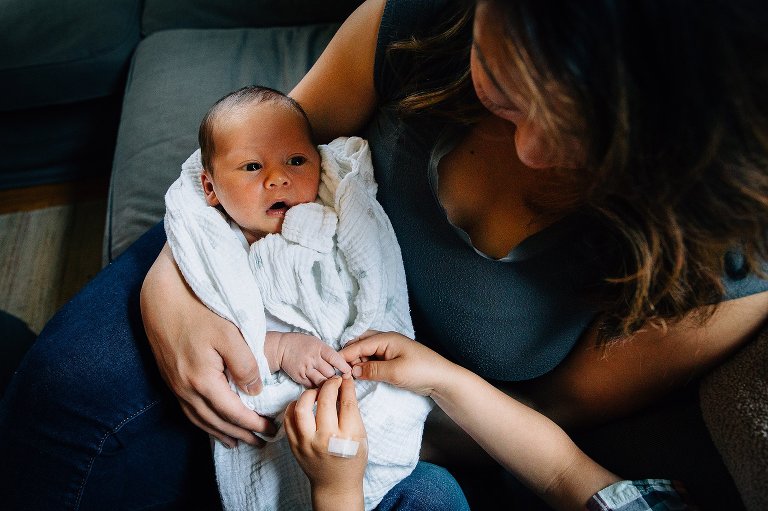 Mother sits on couch holding newborn baby boy. older brother holds his hand looking at his tiny fingers.