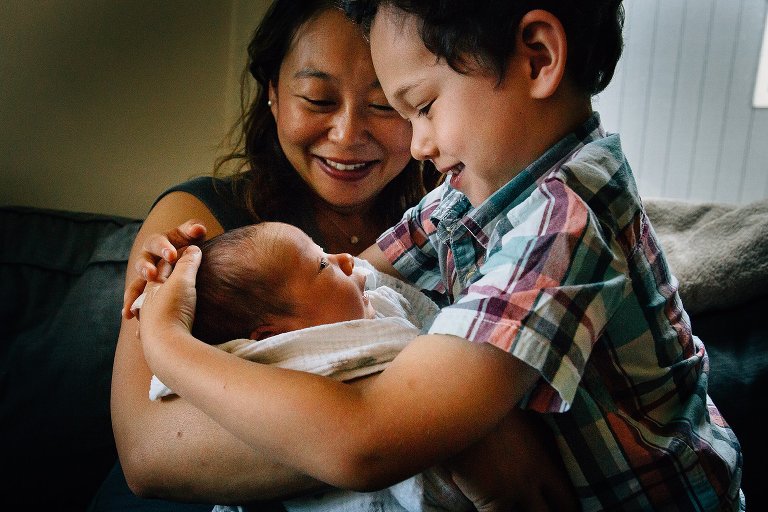 Mother sits on couch holding newborn baby boy as older brother gives him a hug.