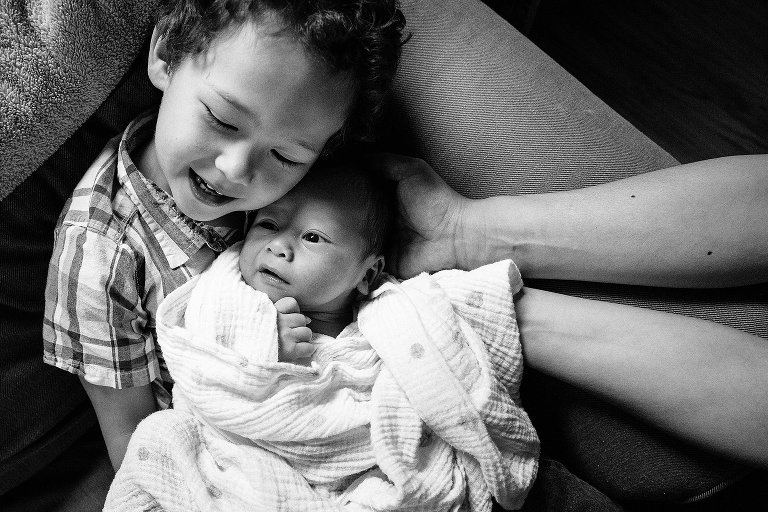 Black and white. Little boy sits on couch as he holds newborn baby brother, mothers hands help support baby's head.
