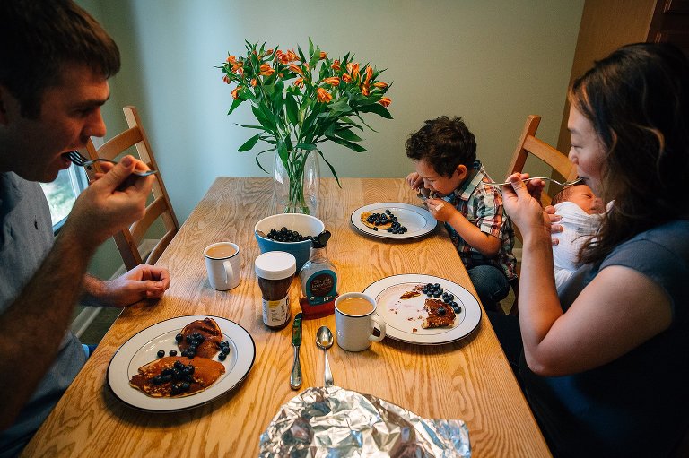 Mother holding newborn baby, Father, and son all sit together at the kitchen table eating blueberries and pancakes for breakfast.
